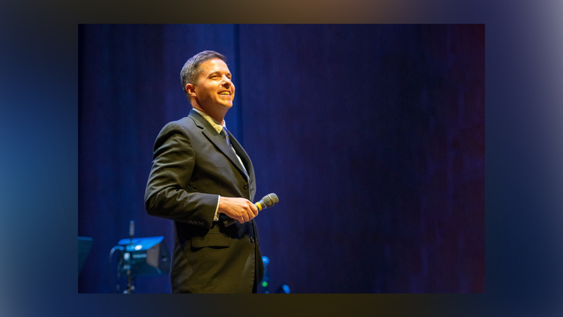 Image of Matt Gibson onstage at the Singletary Center in a black suit against a blue backdrop holding a microphone