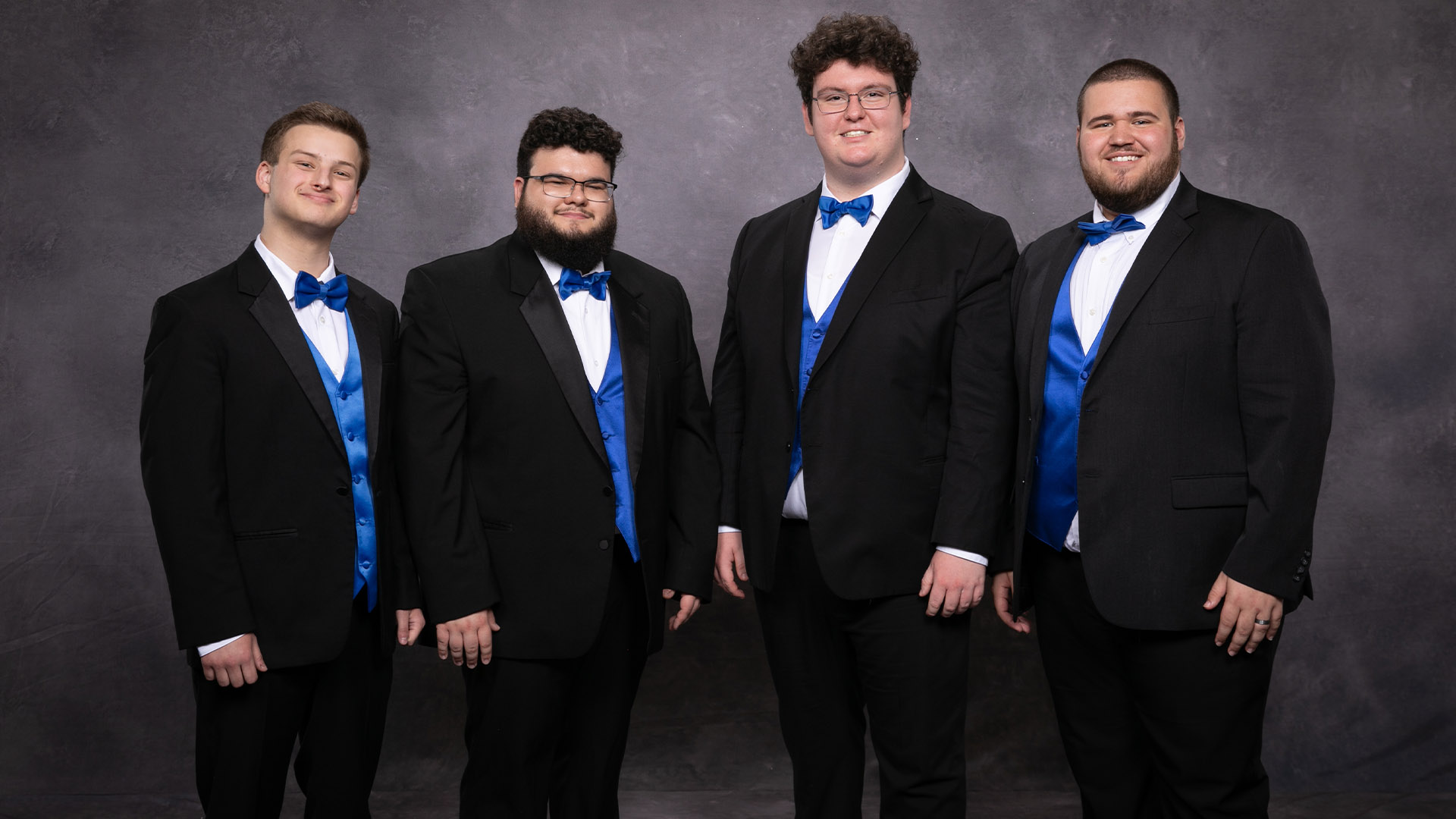 Members of FreshF stand against a grey background wearing tuxedos with blue bowties and vests 