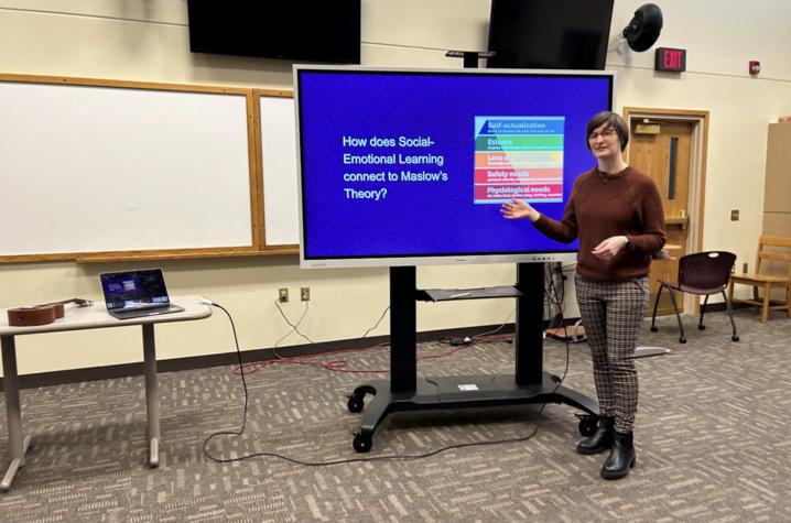 Image of Abigail Van Klompenberg standing in front of a screen that reads "How does social emotional learning connect to Maslow's Theory?"