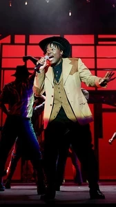 Performers on stage during the 2024 production of Footloose. A man in the spotlight with mic in hand wearing country western attire sings while others dance in the red-lit background.
