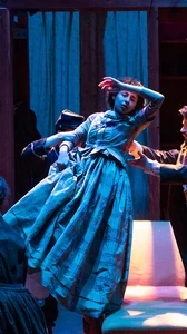 Dramatic blue and gold lighting on a faint woman being gracefully lifted by two men onto a couch while others look on.