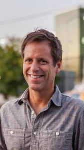 Headshot of Tim Carpenter smiling in denim shirt with blurred cityscape in background.