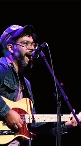 Image of Colin Cutler onstage playing guitar and singing wearing a denim jacket and hat