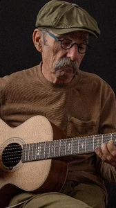 Image of Roy Book Binder seated playing guitar against a black background wearing a tan sweater, black glasses and tan cap