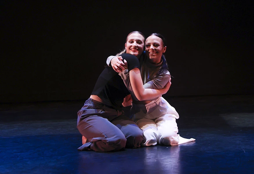 Two dancers on a black stage in dramatic lighting kneeling close to one another and hugging with big smiles.