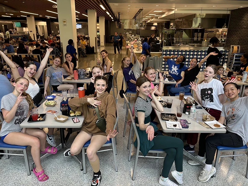 In a busy cafeteria, many dance students sitting at several clustered tables cheerfully pose for the camera over lunch.