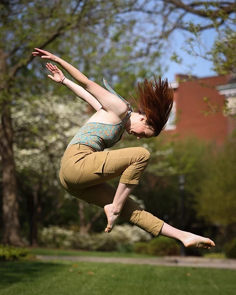 Outdoors on campus, over a pleasant green common area with blooming trees in the background, a dancer with red hair leaps gracefully.