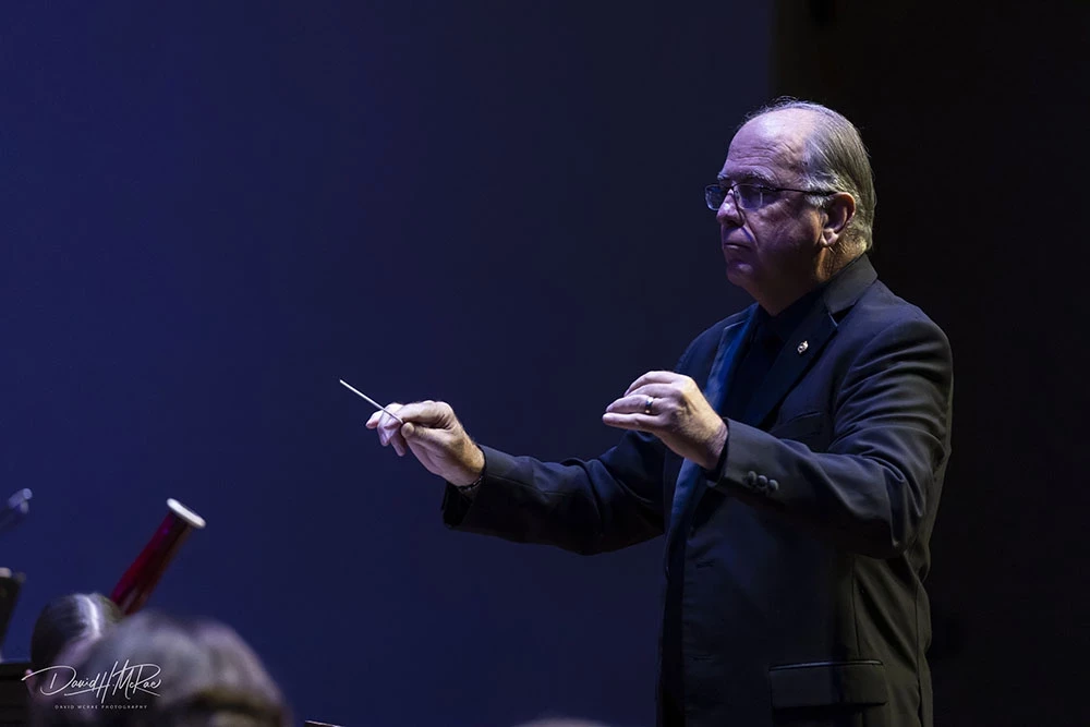 Cody Birdwell holds up both hands, conductor's batton in one hand, while students perform in Singletary with dramatic blue and white lighting.