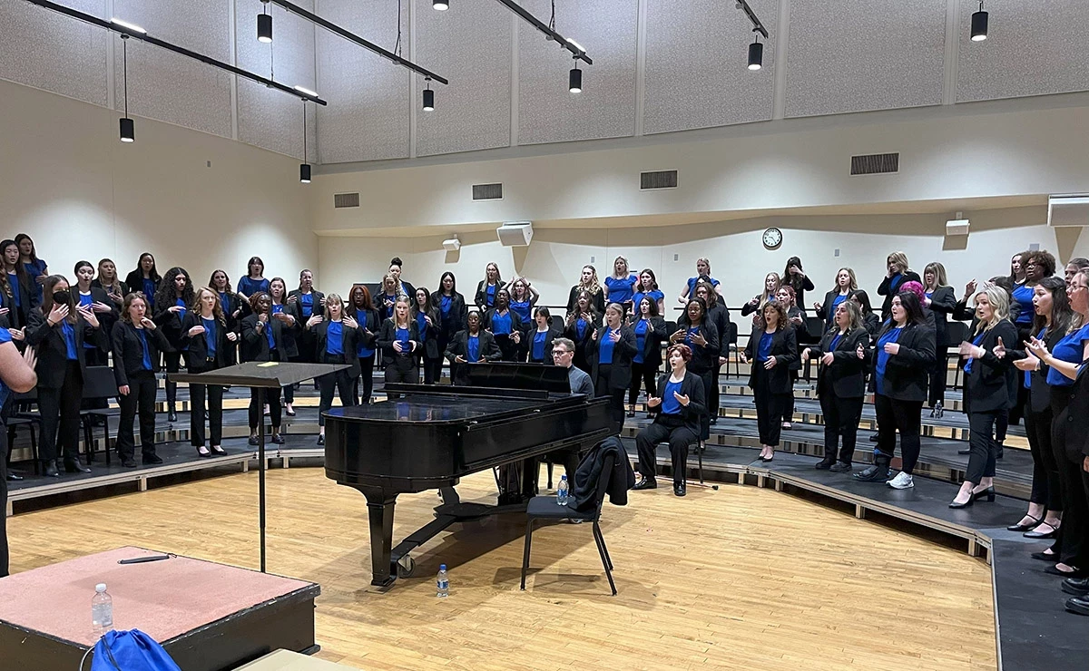 Many singers standing on a three tiered u-shaped riser rehearsing a song with a pianist in the middle of the u-shape accompanying them on a baby grand piano.
