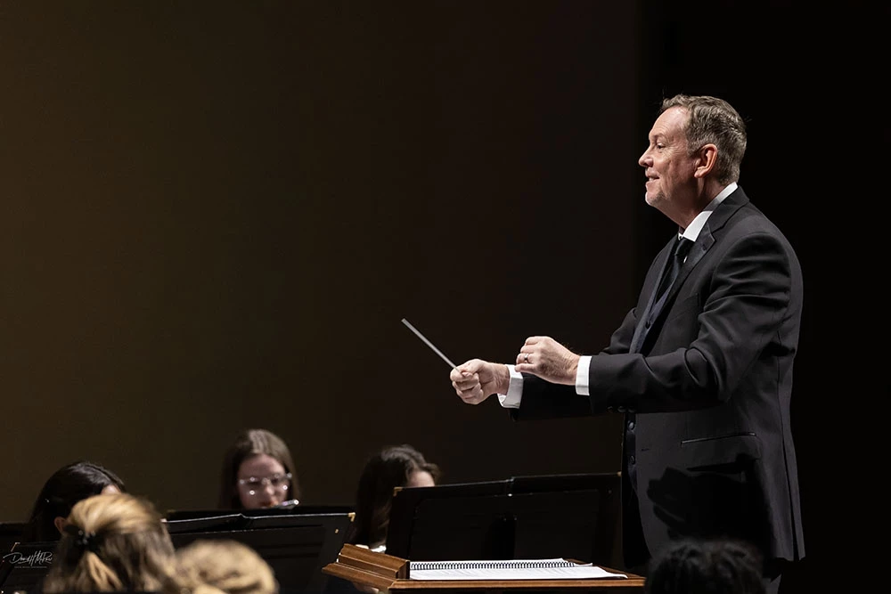 Side view of Brian Froedge as students perform on stage, both hands held out loosely in front of him with one holding up a conductor's batton.