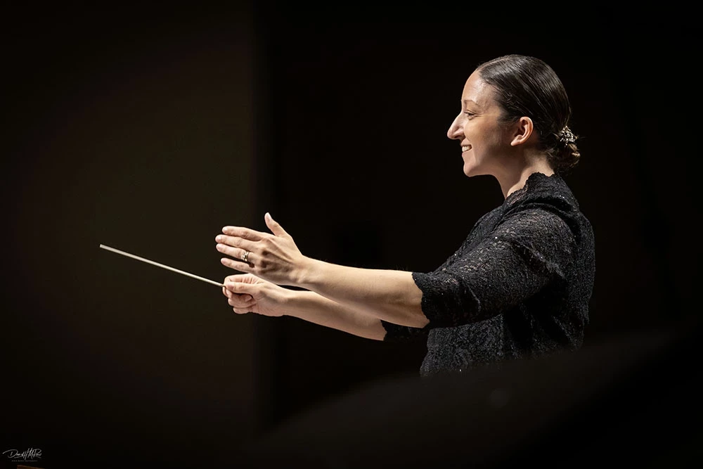 Side view of Shayna Stahl as she smiles at her ensemble, boths hands held out in front of her with one holding a conductor's batton.