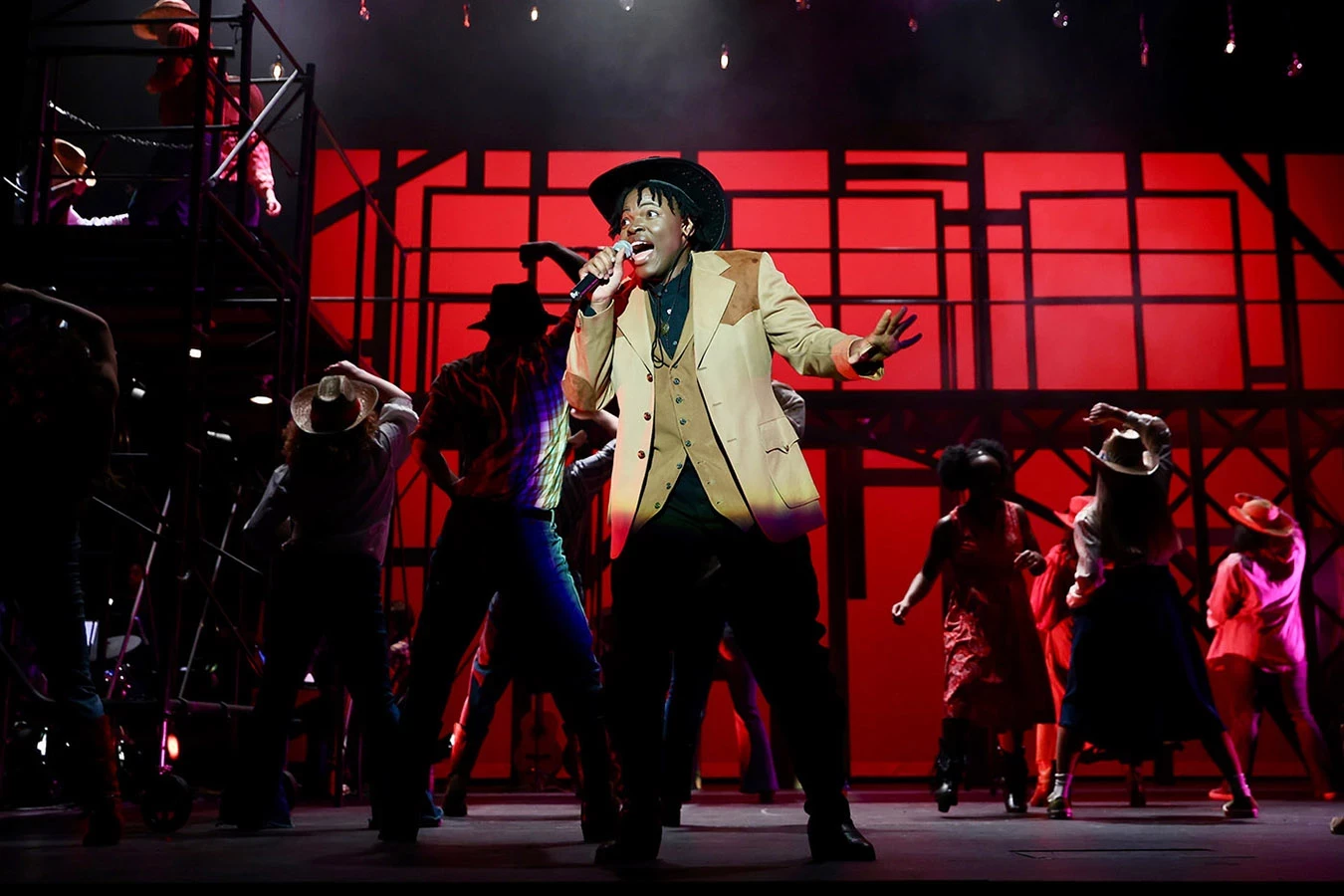 Performers on stage during the 2024 production of Footloose. A man in the spotlight with mic in hand wearing country western attire sings while others dance in the red-lit background.