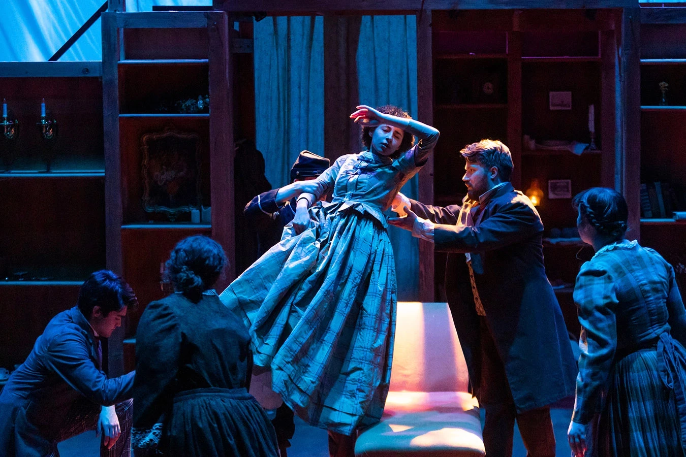 Dramatic blue and gold lighting on a faint woman being gracefully lifted by two men onto a couch while others look on.