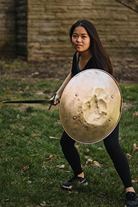 Chelsea Russel in a fighting stance with sword and dented bronze round shield standing on grass in front of a stone building.