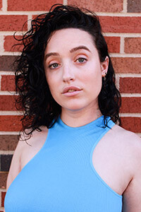 Headshot of Emily Norris in a sky blue tank top standing in front of a brick wall.