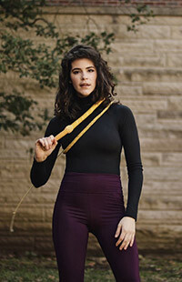 Sam-Claire Beiber stands in front of a stone building with a tan whip looped around her torso.
