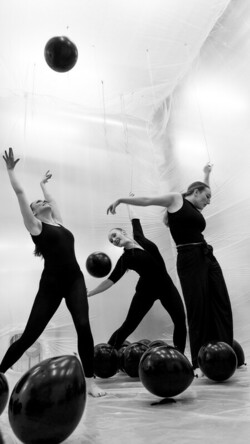 Black and white image of three dancers in a room reaching upwards, with balloons on the floor