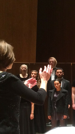 Choristers director in foreground with back to camera conducting members of the UK Choristers onstage, wearing black evening gowns and tuxedos