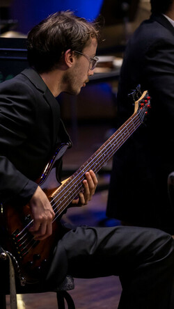 Guitar player sits onstage at the Singletary Center wearing performance blacks playing a guitar and reading music