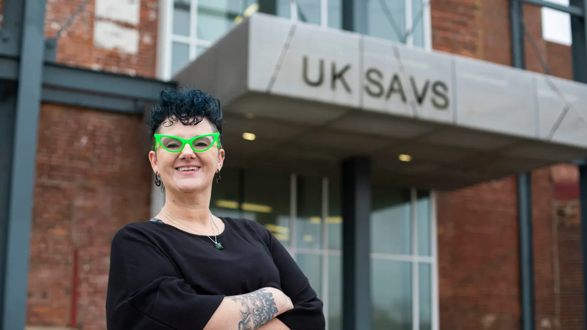 Image of Ambre Armstrong wearing a black top with bright green glasses, standing in front of the SAVS building