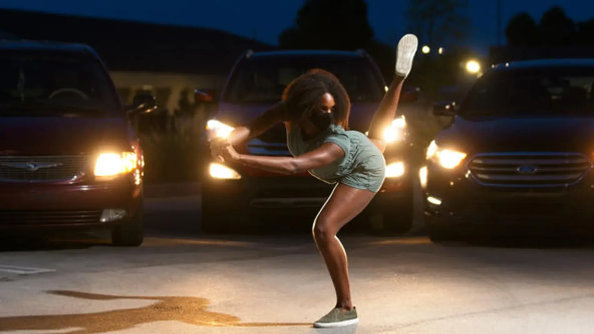 A dancer in a mask performs in parking lot at night surrounded by cars with their headlights illuminating the dancer. The cars are filled with the silhouettes of audience members attending during the pandemic.