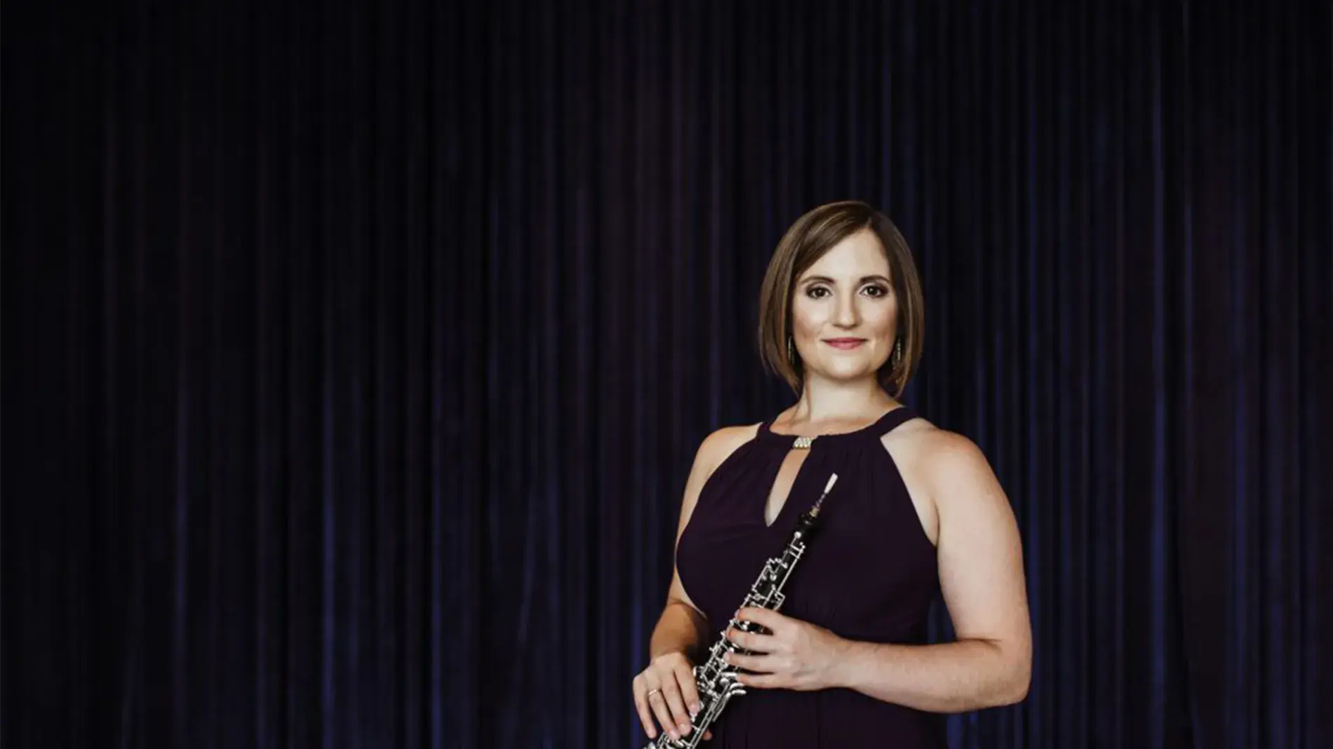 Image of Dr. ToniMarie Marchioni holding oboe, wearing black dress, against black curtain backdrop