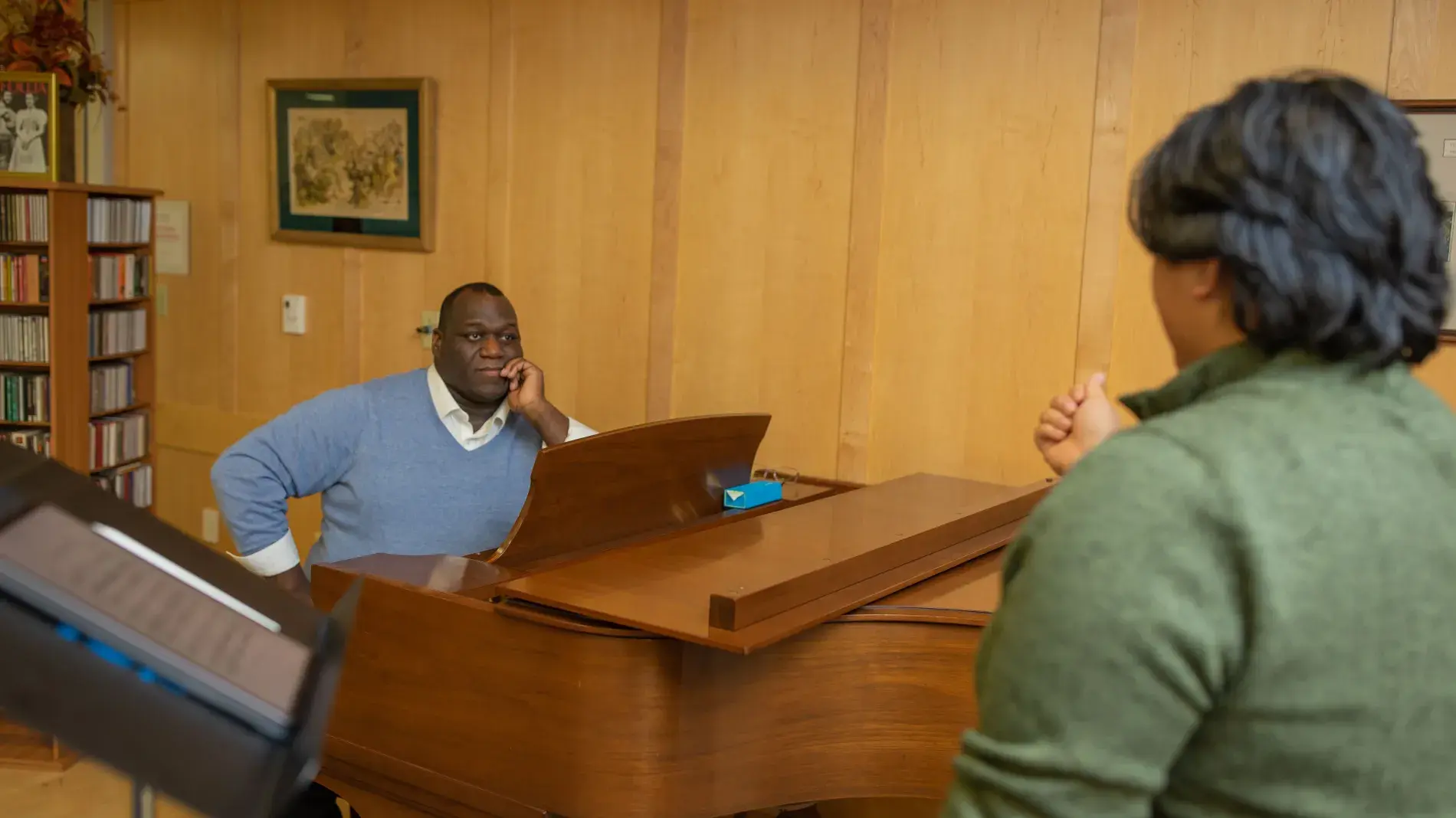 Image of Reggie Smith Jr. seated at a grand piano watching a student in the foreground during a vocal lesson.