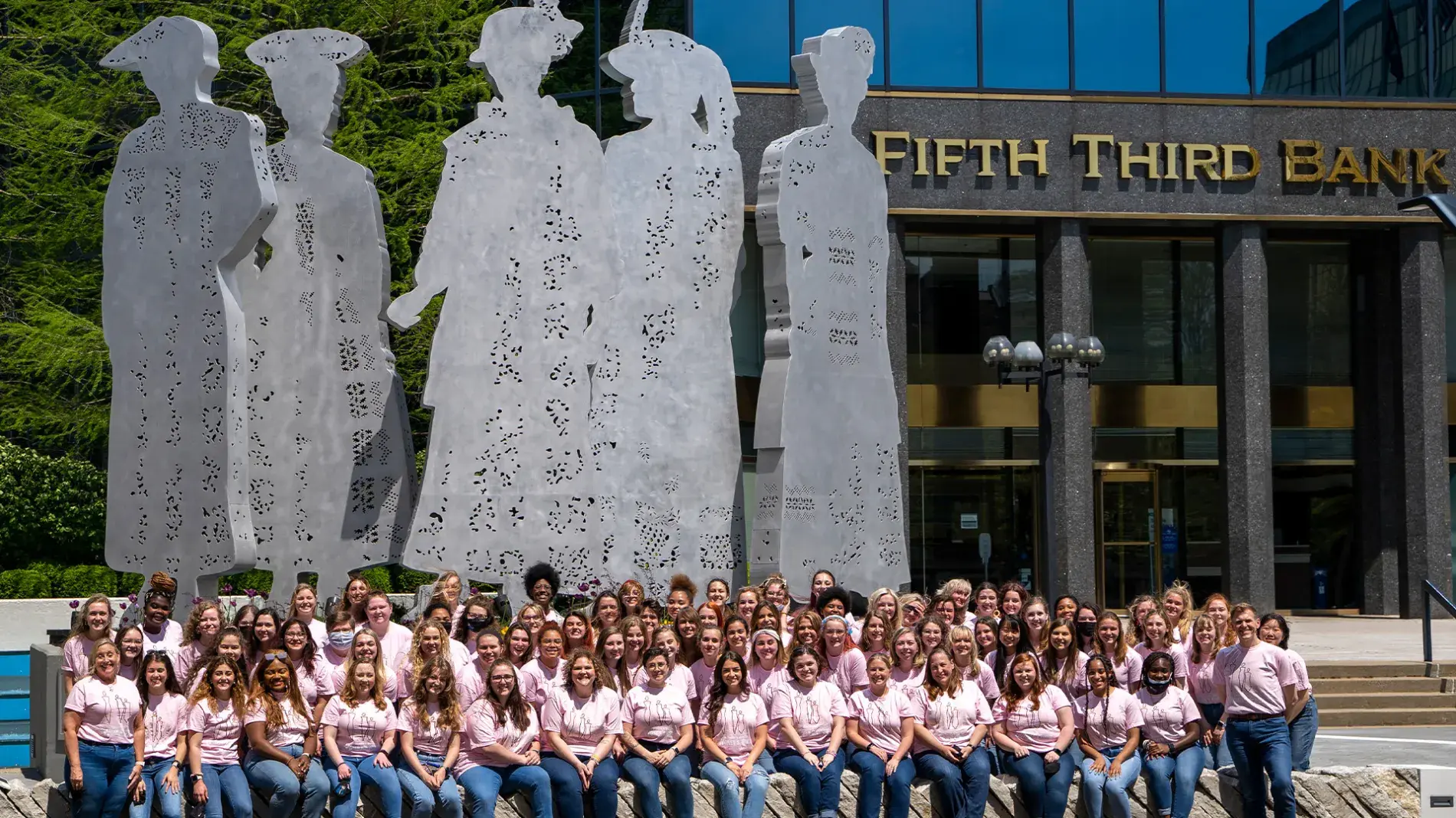 A large group of UK Treble Choir sits outside wearing pink tshirts in front of a large metal statue depicting silhouettes of female suffragettes, in front of the Fifth Third Bank building downtown Lexington  