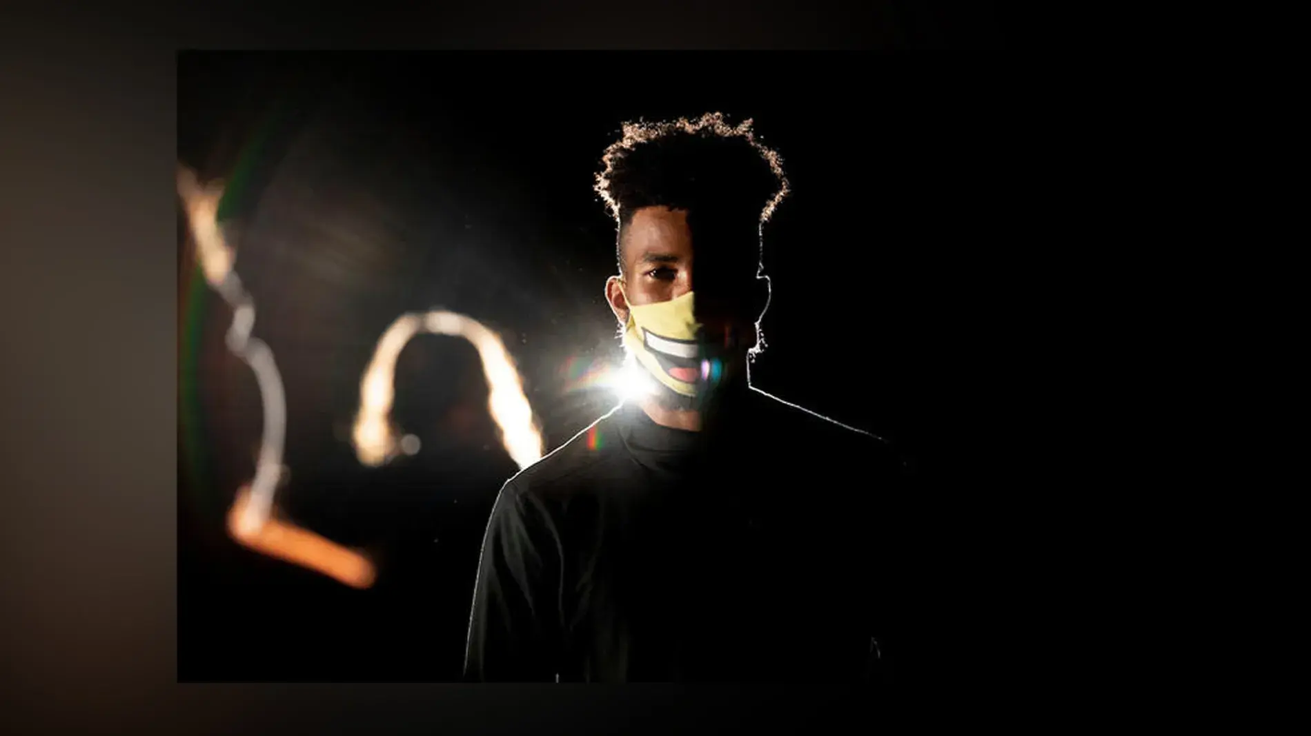 Student wearing white mask and dark shirt faces camera, backlight with other students onstage blurred in background