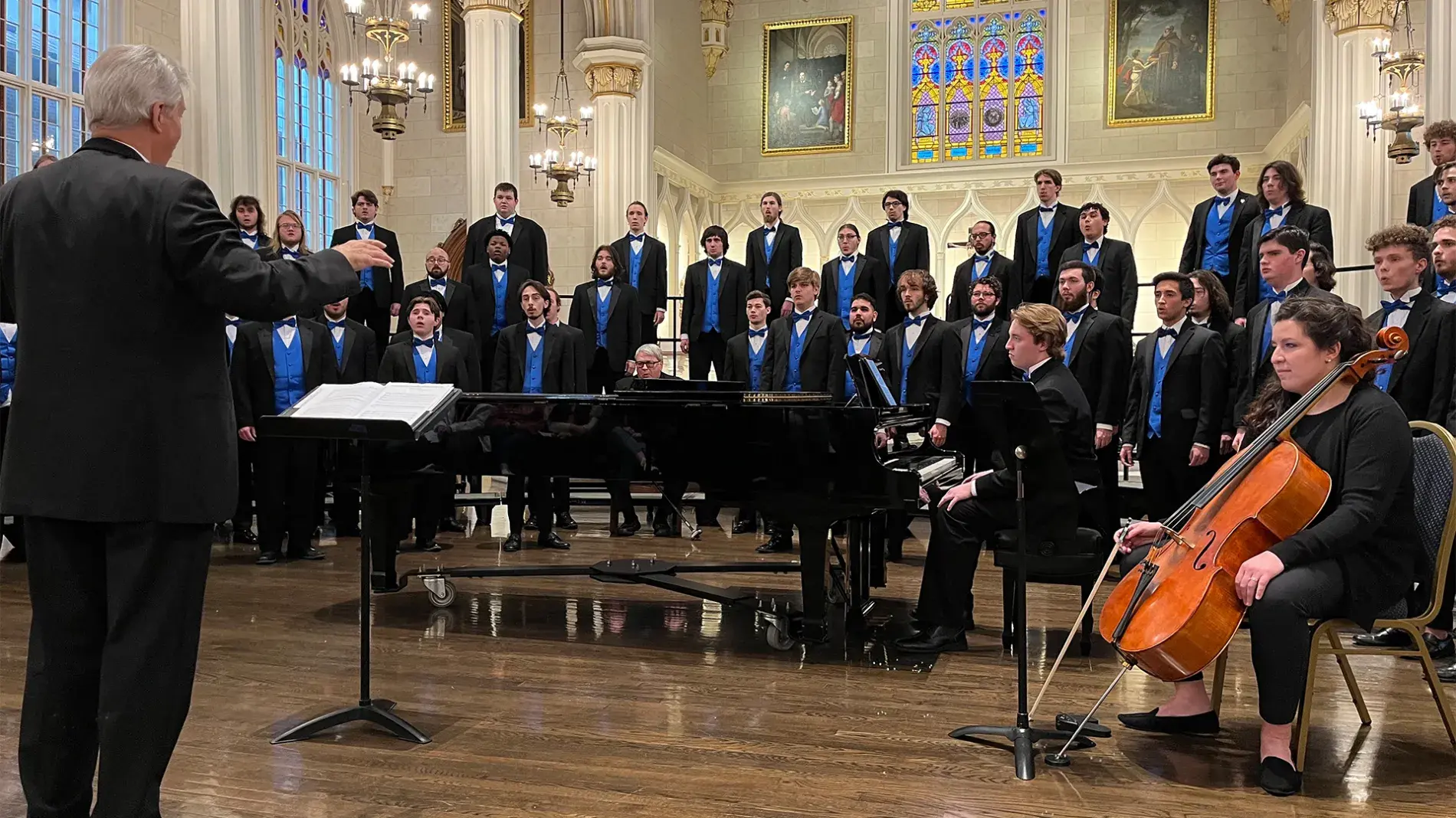 Image of UK Mens Chorus performing, wearing black tuxedos and blue vests, in a cathedral, with musicians and conductor in foreground