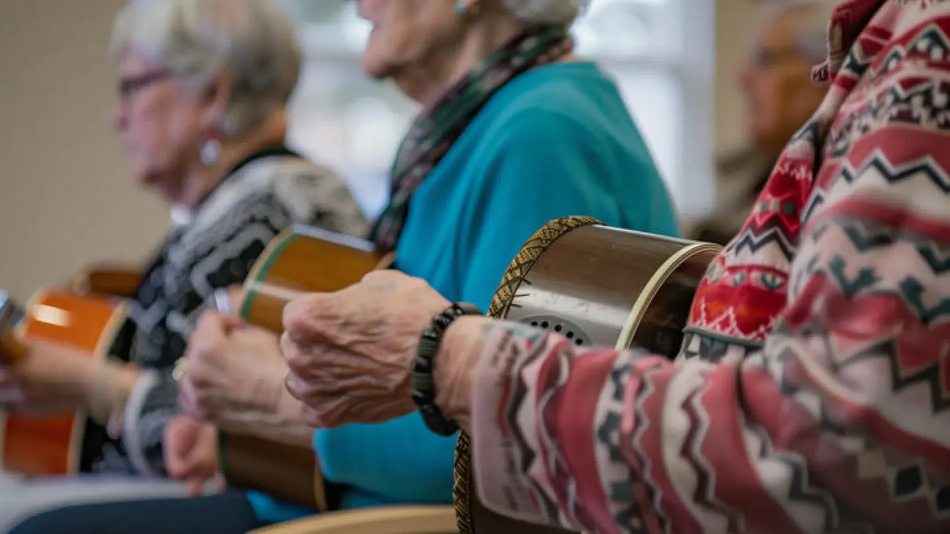 Three older adults sit side by side, playing hand drums, as part of a small group music session or recreational activity in a community or senior center.