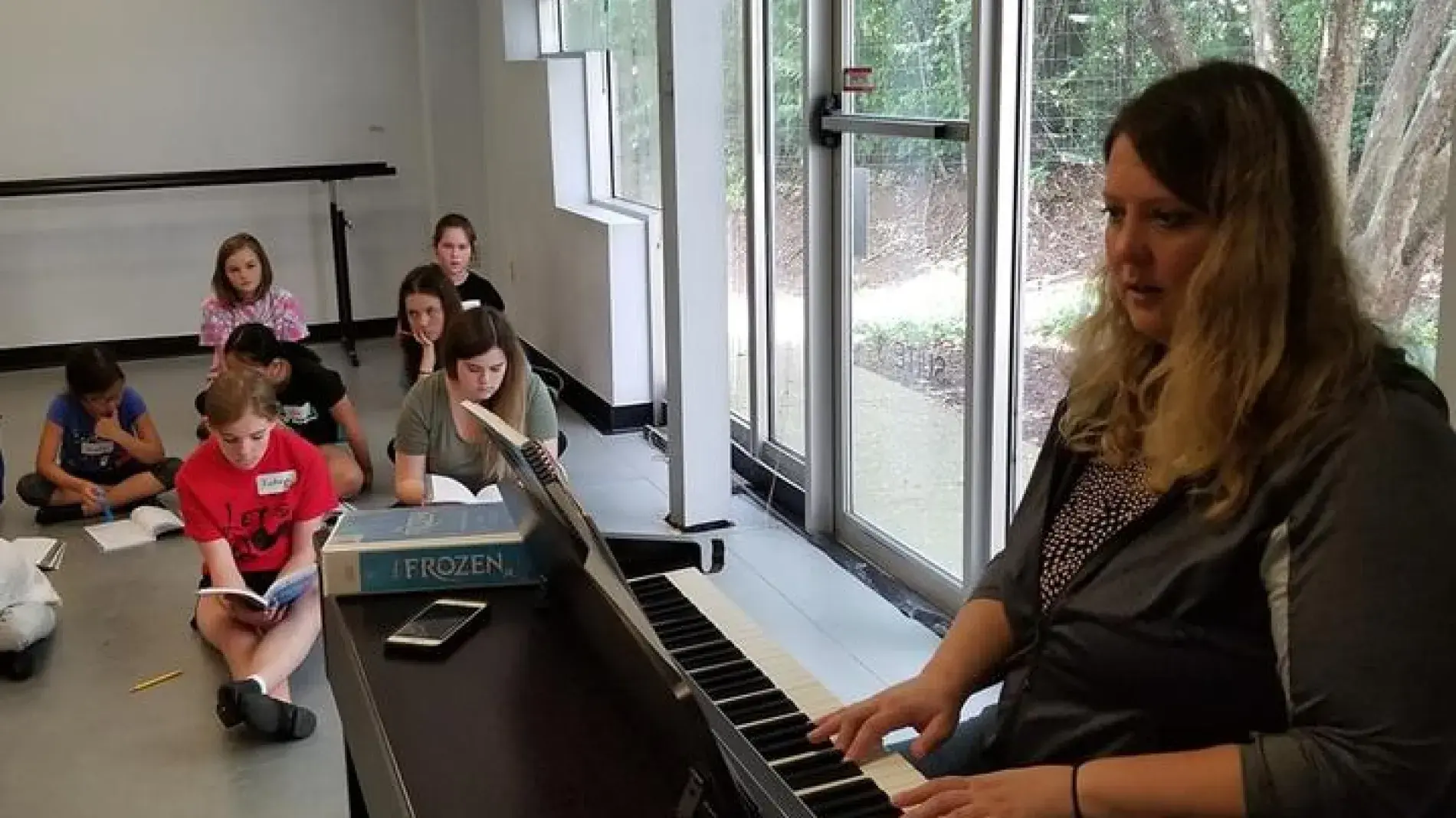 An individual plays an electric keyboard at the front of a bright room while several children sit on the floor in the background near large windows.