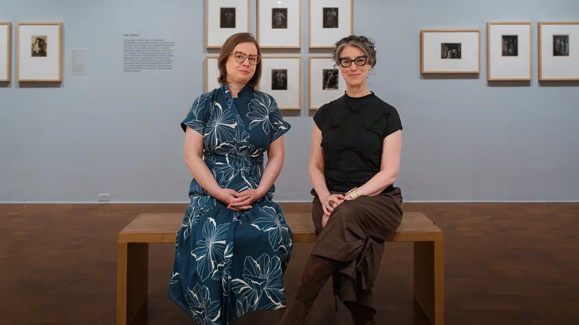Two people sit side by side on a wooden bench in a gallery space, facing the camera. Framed black-and-white photographs line the wall behind them in a neat arrangement, with soft lighting and polished wood floors creating a quiet, museum-like setting.