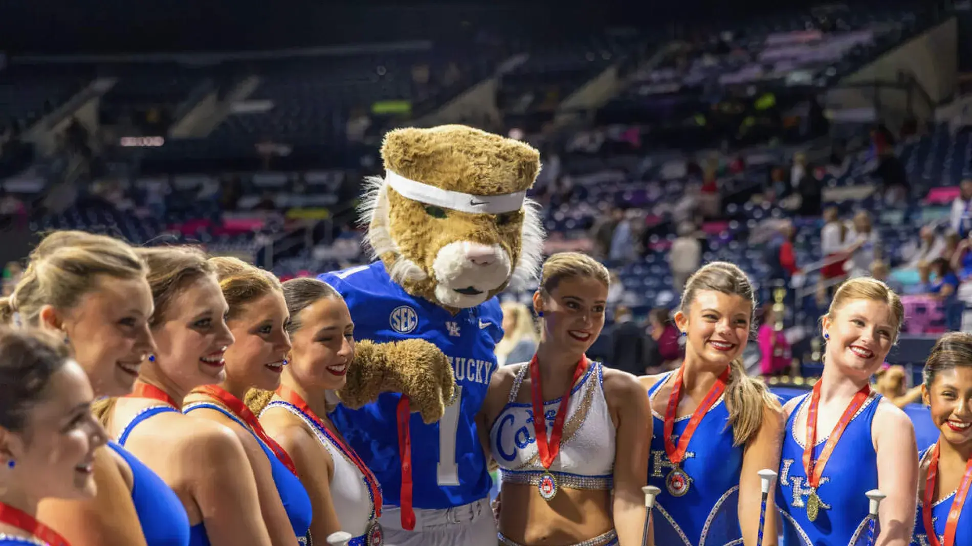 The nine-member team wearing their UK blue uniform with medals around their neck poses for a photo with the Wildcat mascot in front of a crowd.