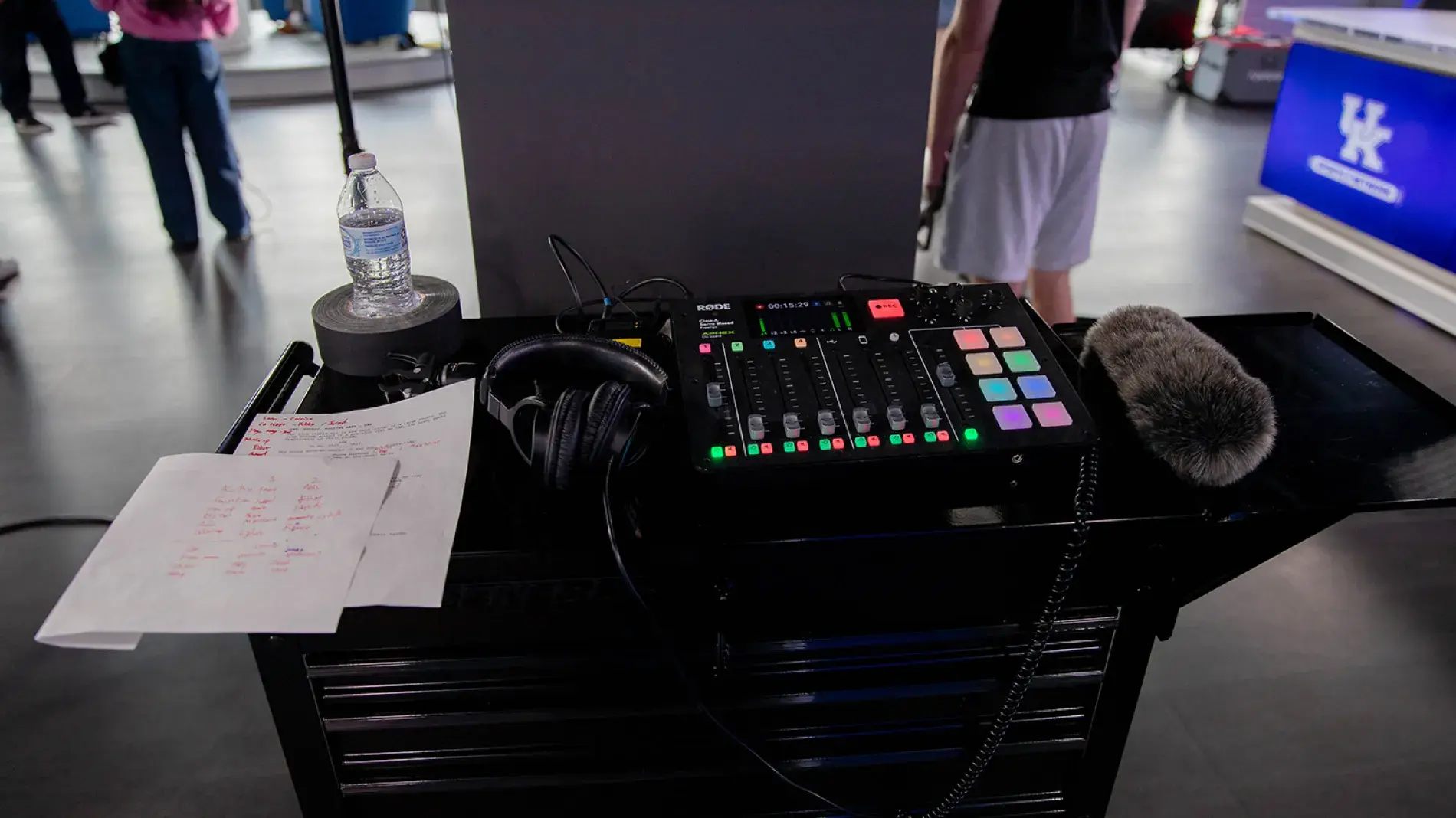 A multichannel mixer sits on a workbench next to a pillar in an open space with several people working in the background. A large mic with a fuzzy wind cover is laying to the right of the mixer. Some headphones and a couple sheets of heavily marked and noted paper lay to the left.