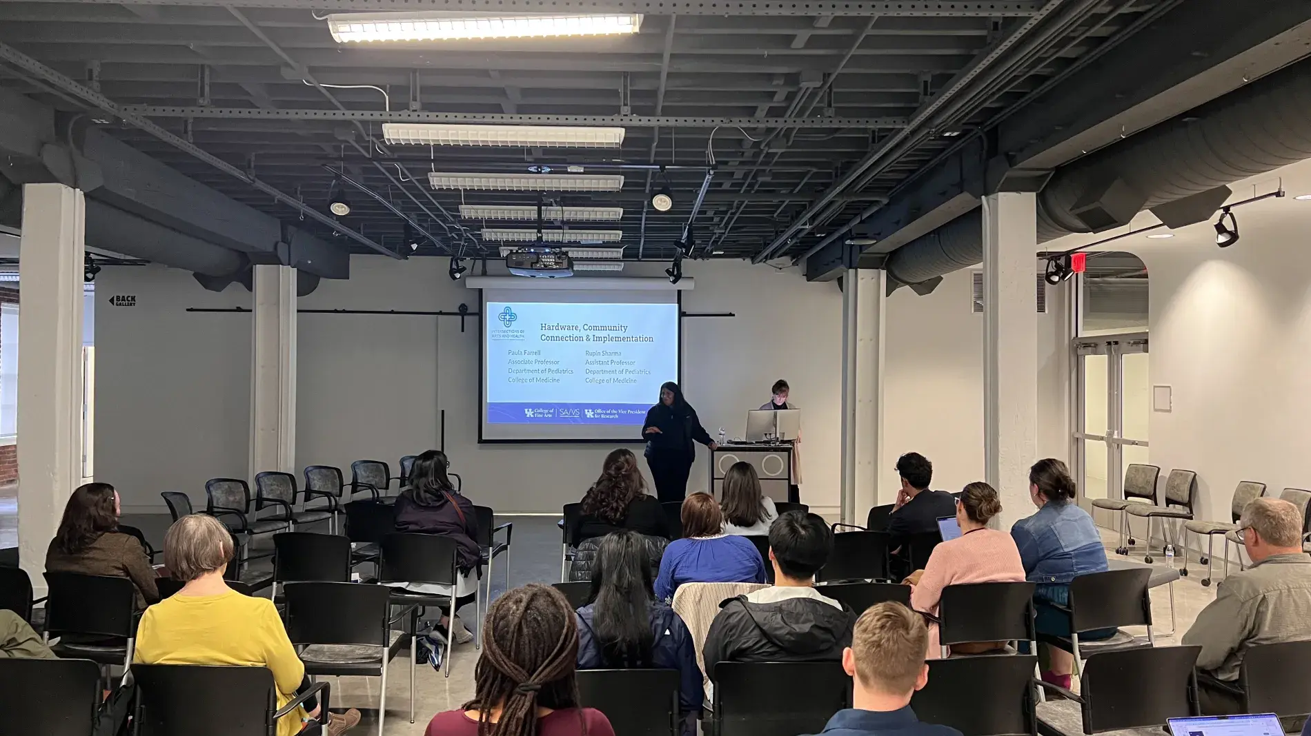 A speaker presents to a seated audience in a loft-style event space with exposed ceiling ductwork. A projection screen displays a slide titled 'Hardware, Community Connection & Implementation' with presenter names Paula Farrell and Rupin Sharma, both from the Department of Pediatrics, College of Medicine, alongside logos for UK College of Fine Arts, SA/VS, and the Office of the Vice President for Research. A presenter stands near a podium with a laptop, addressing about 20 attendees seated in rows of black 