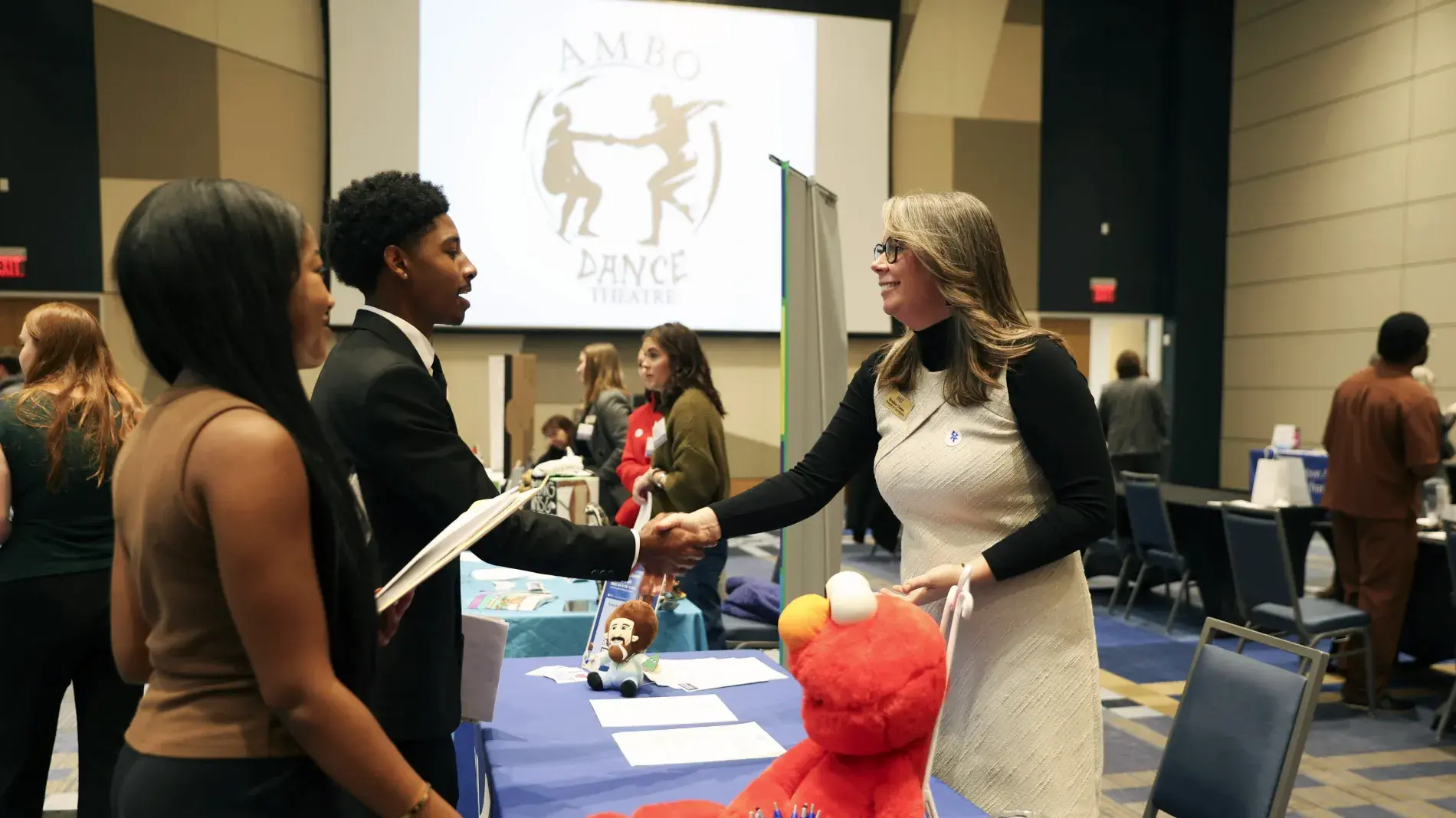 A student in a suit shakes hands with a representative at a career fair table decorated with plush toys, with the AMBO Dance Theatre logo on a screen behind them.