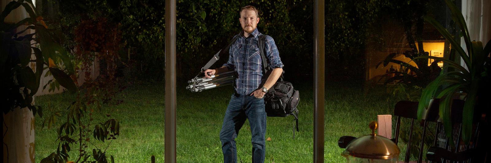 Image of Rob Southard standing outside of window, with his camera around his shoulder at night
