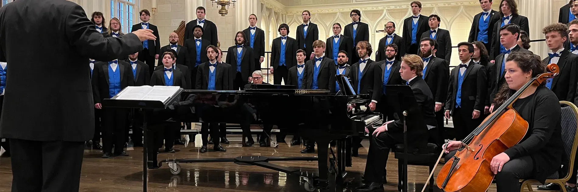Image of UK Mens Chorus performing, wearing black tuxedos and blue vests, in a cathedral, with musicians and conductor in foreground