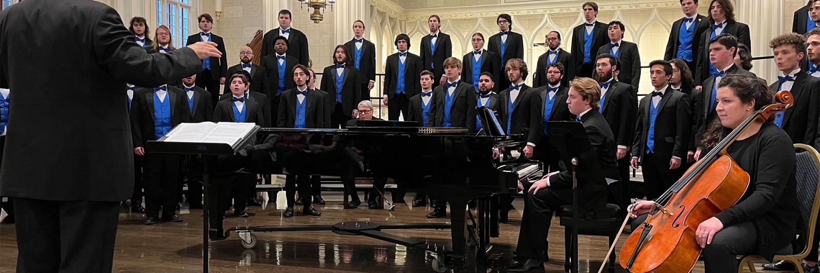 Image of UK Mens Chorus performing, wearing black tuxedos and blue vests, in a cathedral, with musicians and conductor in foreground