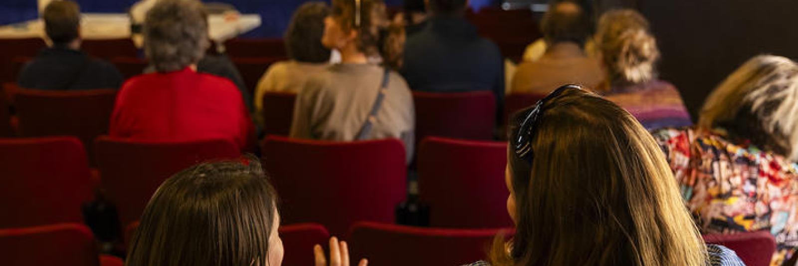 People sit in the audience of a small theatre facing away from camera.