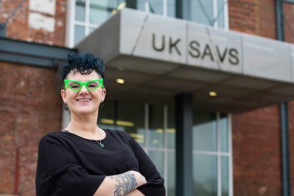Image of Ambre Armstrong wearing a black top with bright green glasses, standing in front of the SAVS building