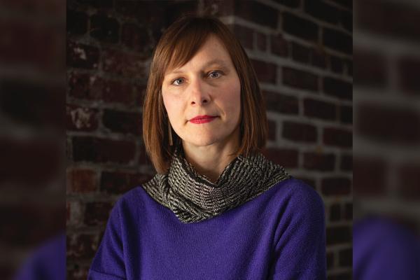 Image of Miriam Kienle, wearing a blue sweater with grey scarf against a brick wall