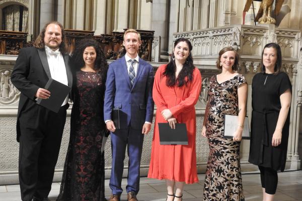 Six vocalists in formal dress stand onstage in front of an ornate backdrop holding their music binders