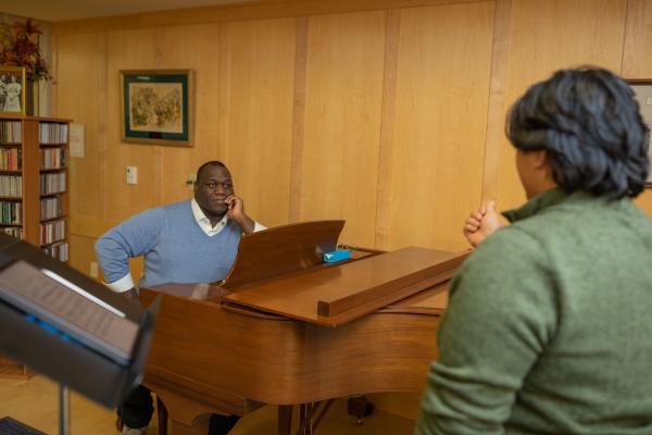 Image of Reggie Smith Jr. seated at a grand piano watching a student in the foreground during a vocal lesson.