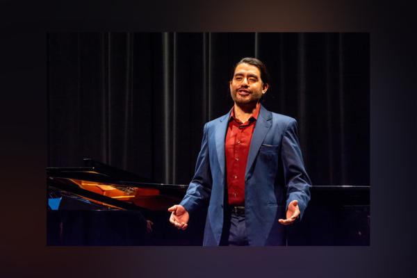 Image of Wagnor Pastor wearing a dark grey suit and red shirt performing onstage in front of a grand piano.