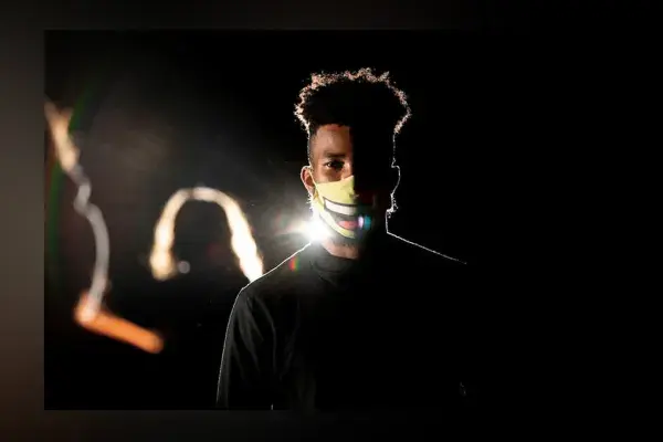 Student wearing white mask and dark shirt faces camera, backlight with other students onstage blurred in background