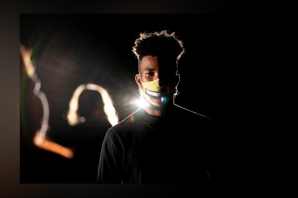 Student wearing white mask and dark shirt faces camera, backlight with other students onstage blurred in background