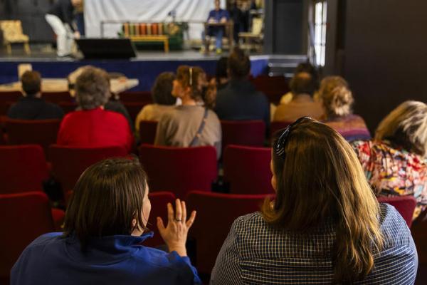 People sit in the audience of a small theatre facing away from camera.