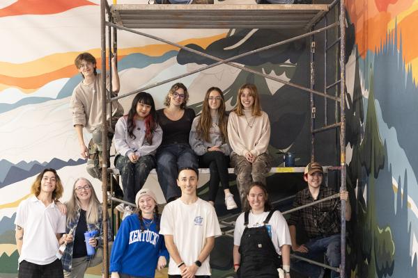 group of students pose in front of large mural in progress on building wall